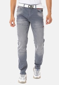 Hellgraue Denim-Jeans in Slim-Fit, mit gesteppten Knieflicken und einem weißen Gürtel. Das Material zeigt leichte Abnutzungserscheinungen mit ausgeblichenen Bereichen.