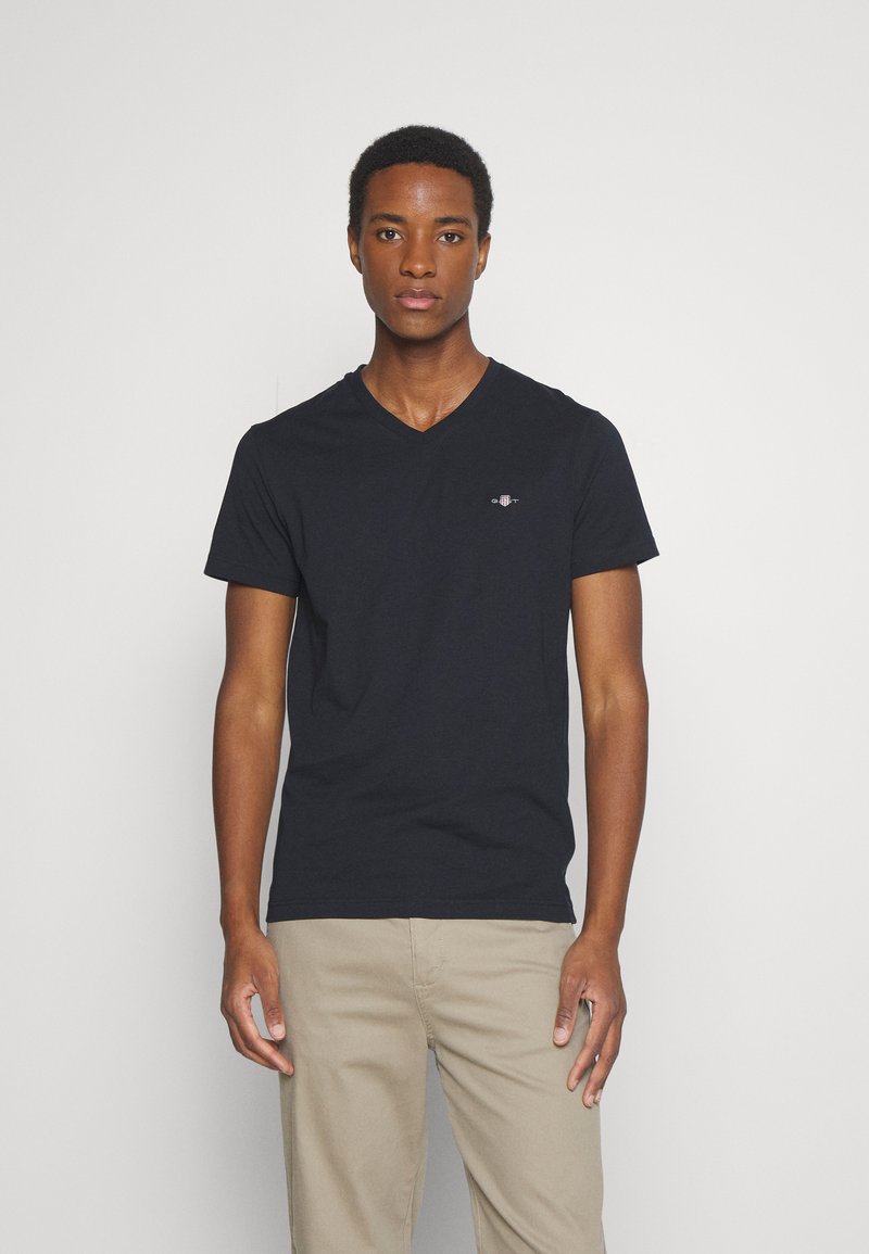 Camiseta de algodón negra con cuello en V y mangas cortas, con un pequeño logo bordado en el lado izquierdo del pecho. Textura suave, fit casual.