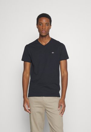 Jeune homme debout de face, portant un t-shirt noir encolure en V et un pantalon beige sur un fond clair uni.