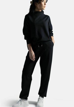 Nainen seisoo käsi taskussa, yllään musta collegepaita ja housut, valkoiset lenkkarit, katsoo sivulle yksinkertaisen taustan edessä.