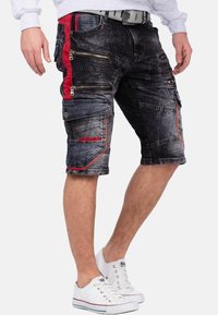 Zwarte denimshorts met rode accenten aan de zijkant, meerdere met rits afsluitbare zakken, een relaxte pasvorm en een rafelrand. Gestyled met witte sneakers.