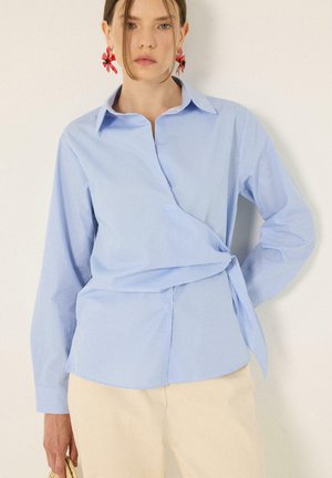 Femme portant une chemise portefeuille à fines rayures bleu clair, un pantalon crème et des boucles d'oreilles rouges en forme de fleur, debout devant un fond uni clair.