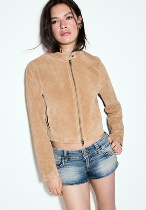Femme aux cheveux foncés portant une veste en suède marron clair à fermeture éclair et un short en denim bleu, debout devant un fond blanc uni.