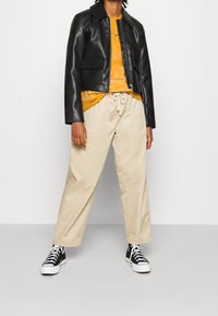 Giacca di pelle nera con colletto, t-shirt arancione sotto, pantaloni beige a gamba larga e sneakers nere con suola bianca.