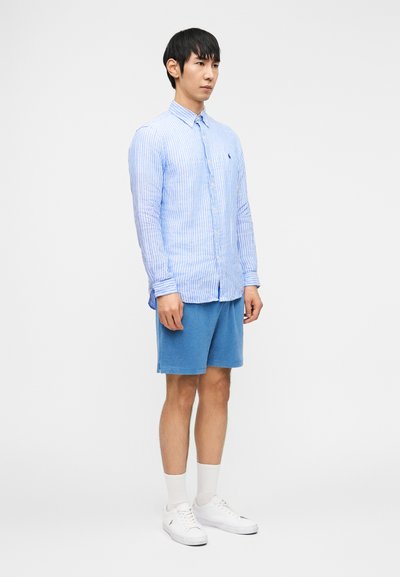 Άνδρας που στέκεται φορώντας ένα ανοιχτό μπλε ριγωτό πουκάμισο με κουμπιά, μπλε σορτς, λευκές κάλτσες και λευκά αθλητικά παπούτσια σε απλό λευκό φόντο.