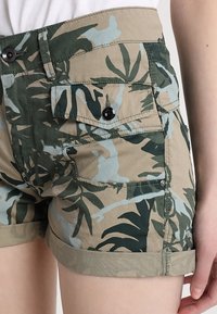 Khaaki kraťasy s camouflage vzorem v zelené a modré, obsahující dvě přední kapsy, ohrnuté lemy a černé knoflíkové detaily.