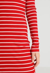 Punainen ribbinen pitkähiha dressi, jossa on valkoisia vaakaraitoja. Ominaisuuksiin kuuluu säädettävä malli ja teksturoitu kangas, joka korostaa puhtaita linjoja ja venyvyyttä.