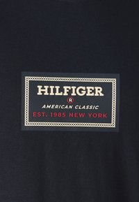 Camiseta de color azul marino oscuro con una etiqueta rectangular de tela. La etiqueta muestra "HILFIGER" en blanco, "AMERICAN CLASSIC" en dorado y "EST. 1985 NEW YORK" en rojo.