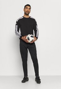 Svart langermet treningstrøye med hvite striper på ermene, svarte tapered bukser, svarte fotballsko, holder en mønstrete fotball.