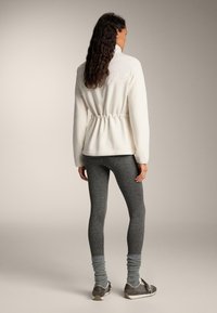 Jachetă albă din fleece cu talie strânsă, asortată cu colanți gri. Ținuta are un guler înalt și un material texturat pentru căldură.