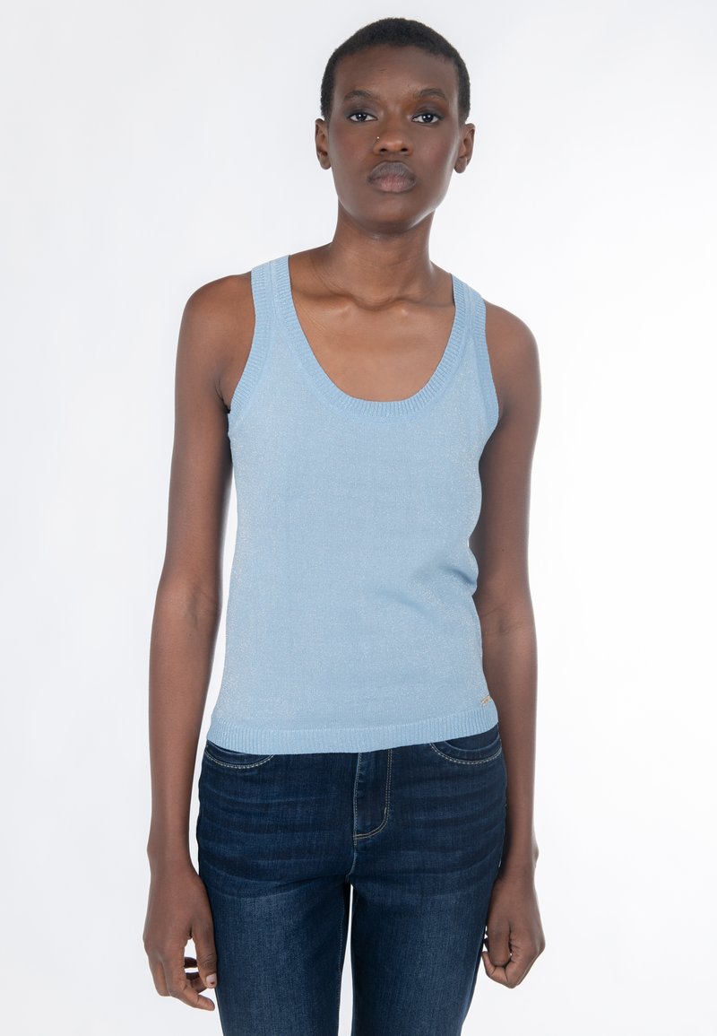 Mujer de pie contra fondo blanco, vistiendo una camiseta de punto sin mangas de color azul claro y jeans azul oscuro, mirando al frente con expresión neutral.