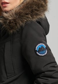 Black puffer jacket featuring a fur-lined hood, a round blue and black embroidered patch on the left sleeve, and quilted texture on the fabric.