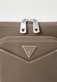 Brun teksturert stoffkelt med sølvhardware, to glidelåslommer og en trekantslogo med "GUESS USA."