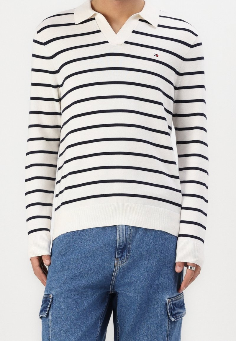 Personne portant un polo blanc à manches longues à rayures horizontales noires et un jean cargo bleu, tenant un téléphone dans la main droite.