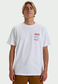 Young man with blond hair wearing a white T-shirt with small red and blue "FINZZA Billabong Co." logo on chest, brown pants, standing against plain background.