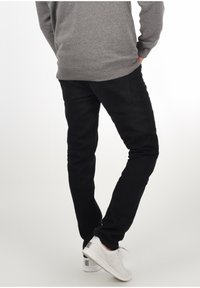 Jeans neri slim fit con cinque tasche, indossati con un maglione grigio e scarpe da ginnastica bianche. I jeans hanno una texture liscia e una leggera elasticità.