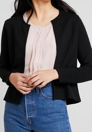 Femme portant une veste noire texturée ouverte sur le devant, un chemisier beige plissé et un jean bleu taille haute, mains jointes devant.