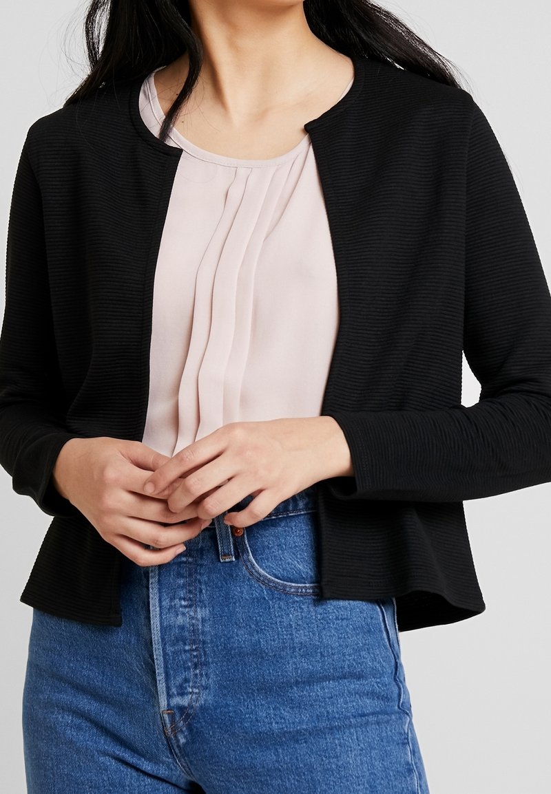 Femme portant une veste noire texturée ouverte sur le devant, un chemisier beige plissé et un jean bleu taille haute, mains jointes devant.