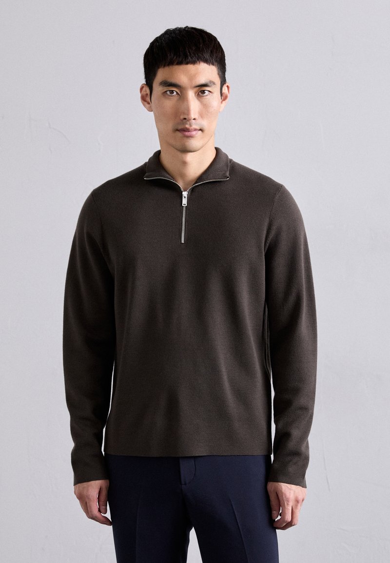Uomo con capelli neri corti che indossa un maglione zip-up marrone scuro e pantaloni blu navy, in piedi contro un muro grigio chiaro uniforme.
