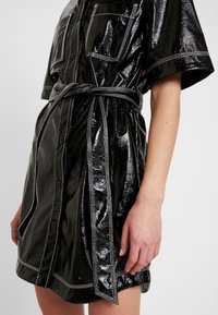 Svart kort knäppklänning i glänsande material, med kontrasterande vit sömnad, två framfickor och ett knytbälte i midjan.