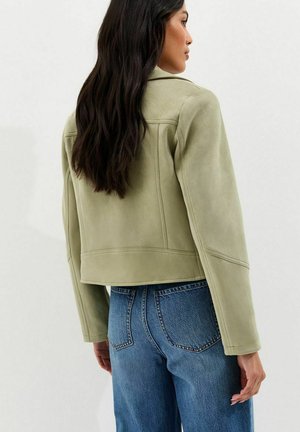 Vrouw met lang donker haar, gekleed in een lichtgroene jas en blauwe spijkerbroek, van achteren gezien tegen een effen achtergrond.