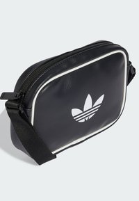 Čierna syntetická crossbody taška s bielym lemovaním a veľkým bielym logom Adidas. Obsahuje zips a nastaviteľný popruh.