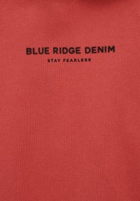 Camiseta de mezcla de algodón en color coral con texto impreso en negro: "BLUE RIDGE DENIM" y "STAY FEARLESS". Textura suave, diseño de cuello redondo.