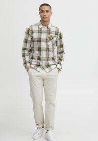 Camisa de cuadros en blanco, verde y amarillo, con un bolsillo en el pecho. Combinada con pantalones beiges y zapatillas blancas. Ajuste casual.