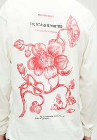 Weißes Langarmhemd mit großem rotem Blumenmuster und dem Text "DIE WELT WARTE AUF DICH" sowie motivierenden Zitaten auf der Rückseite.