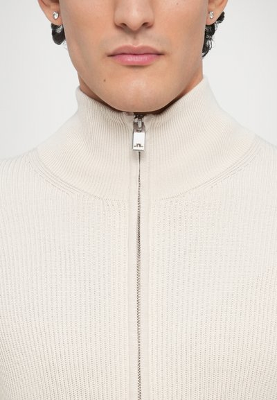 Πλεκτό μπεζ πουλόβερ με φερμουάρ και ψηλό γιακά, με ραμμική υφή και λεπτομέρεια ασημένιου φερμουάρ στον λαιμό. Απλός και εφαρμοστός σχεδιασμός.