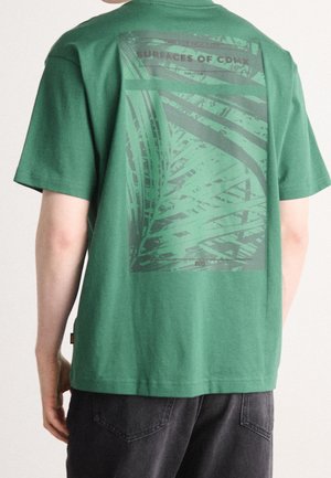 Person, die ein grünes T-Shirt mit einer dunklen abstrakten rechteckigen Grafik und dem Text „SURFACES OF CDMX“ auf dem Rücken trägt.