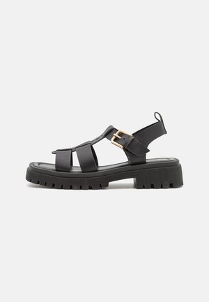 Sort sandal med chunky sål, tre remme og guldlås på siden, åben tå og hæl samt trækflap bagpå.
