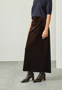 Persona che indossa una lunga gonna di velluto marrone scuro, una maglietta a maniche corte blu navy e stivaletti con tacco alla caviglia in pelle di coccodrillo marrone scuro.