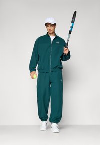 Ensemble de survêtement vert avec une veste zippée et des poignets élastiques, associé à un pantalon assorti. Le modèle tient une raquette de tennis et une balle, portant une casquette blanche.