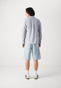 Blå og hvitstripet langermet skjorte med knapper ved mansjettene, kombinert med lyseblå denimshorts og brune slip-on sko med hvite sokker.