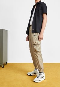 Černá krátká rukávová košile, béžové cargo kalhoty s kapesními zipy a šedobílé tenisky Nike. Hladký materiál, pohodlný střih a viditelné logo.