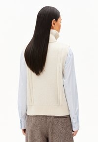 Crème-kleurige gebreide mouwloze coltrui met gestructureerde patronen en een ribgebreide zoom, gedragen over een lichtblauw langemouwen shirt.