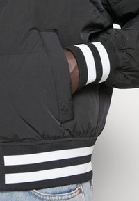 Chaqueta bomber negra de nailon con un bolsillo con cremallera, puños acanalados con rayas blancas y un dobladillo acanalado a juego.