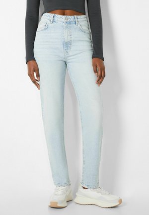 Person trägt hellblaue High-Waist-Jeans, schwarzen langärmeligen Crop-Top und weiße Sneakers, steht vor einem einfarbigen weißen Hintergrund.