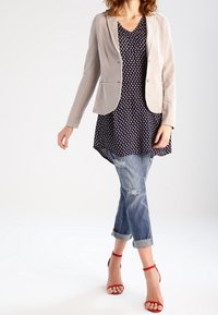 Lichtbeige blazer over een marineblauwe tuniek met stippen, gecombineerd met versleten, opgerolde jeans en rode gekruiste hakken. Focus op gestructureerde stof en pasvorm.