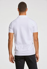 Valkoinen lyhythihainen poolopaita pehmeällä tekstuurilla ja kauluksella, nähtynä takaa, yhdistettynä tummiin housuihin.