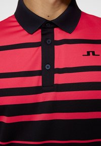 Czerwono-czarny polo w paski z czarnym kołnierzykiem. Posiada trzy czarne guziki i małe logo po lewej stronie. Gładka tekstura materiału.