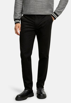 Black chinos with a smooth finish, standard fit, front pockets, worn with black leather boots. The model is standing with hands in pockets.
