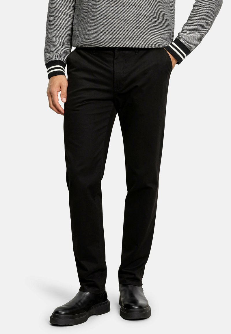 Schwarze Chinos mit glatter Oberfläche, normaler Schnitt, mit Fronttaschen, getragen mit schwarzen Lederstiefeln. Das Modell steht mit den Händen in den Taschen.
