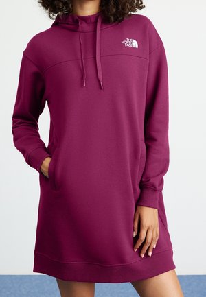 Kobieta ubrana w sukienkę-bluzę o długich rękawach w kolorze śliwkowym z przednimi kieszeniami i logo The North Face na piersi.