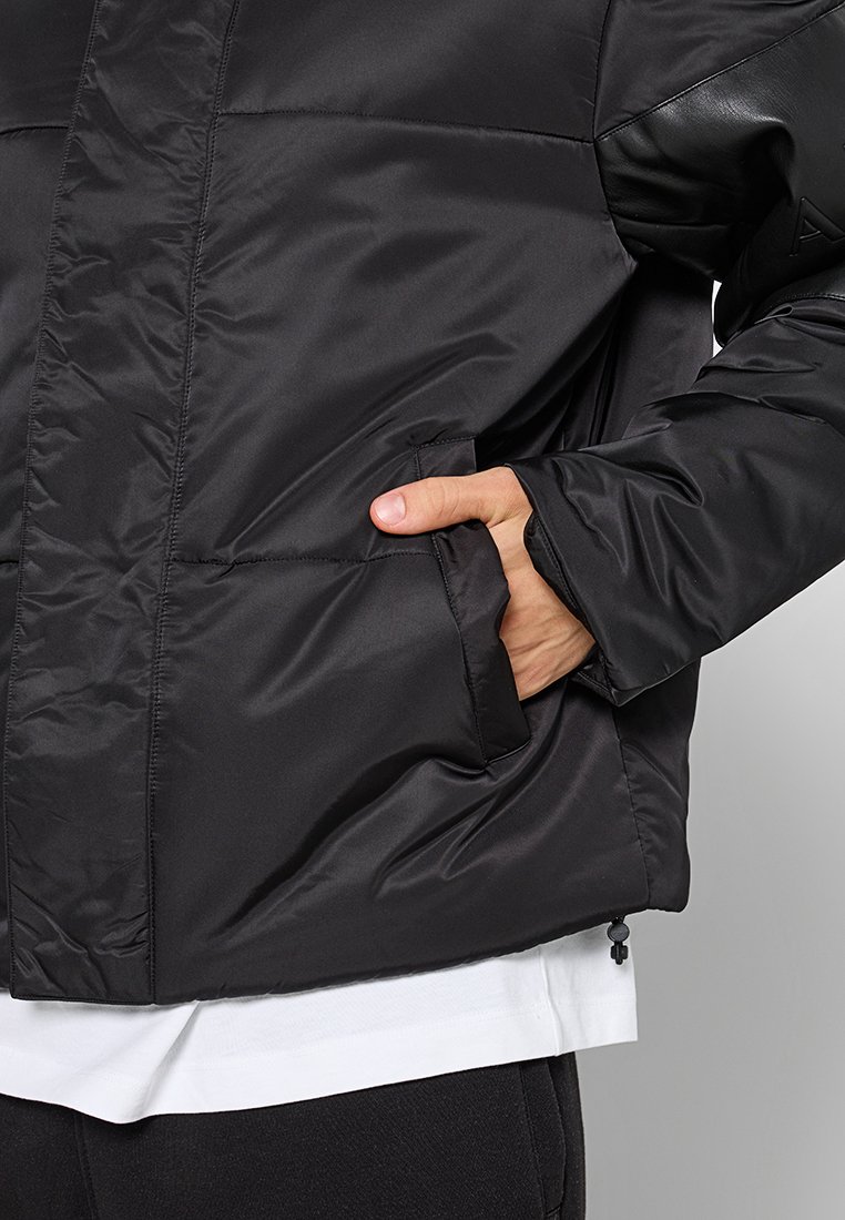 Giacca nera imbottita con tessuto liscio e lucido, dotata di tasca con cerniera, polsini elasticizzati e uno strato interno bianco che sbuca dal fondo.
