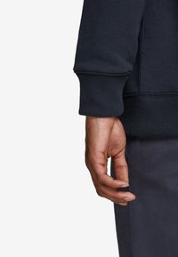 Sudadera de un oscuro color marino con el brazo mostrando un puño acanalado y una textura suave. La mano está posicionada contra una tela gris, mostrando un agarre relajado.