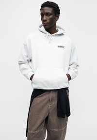 Ανοιχτό γκρι φούτερ με κορδόνι και τσέπη μπροστά, με μαύρο λογότυπο "OBEY". Φορεμένο με καφέ παντελόνι και μαύρο πουκάμισο δεμένο στη μέση.