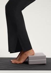 Siv blok za jogo leži na črni teksturirani podlogi, na vrhu katere je bosa noga. Vidne so ohlapne črne hlače s stranskim razporkom.