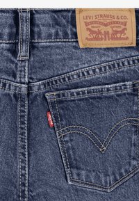 Mørkeblå denimjeans med et lærmerke med preget logo, gule sømmer og en liten rød etikett på baklommen.